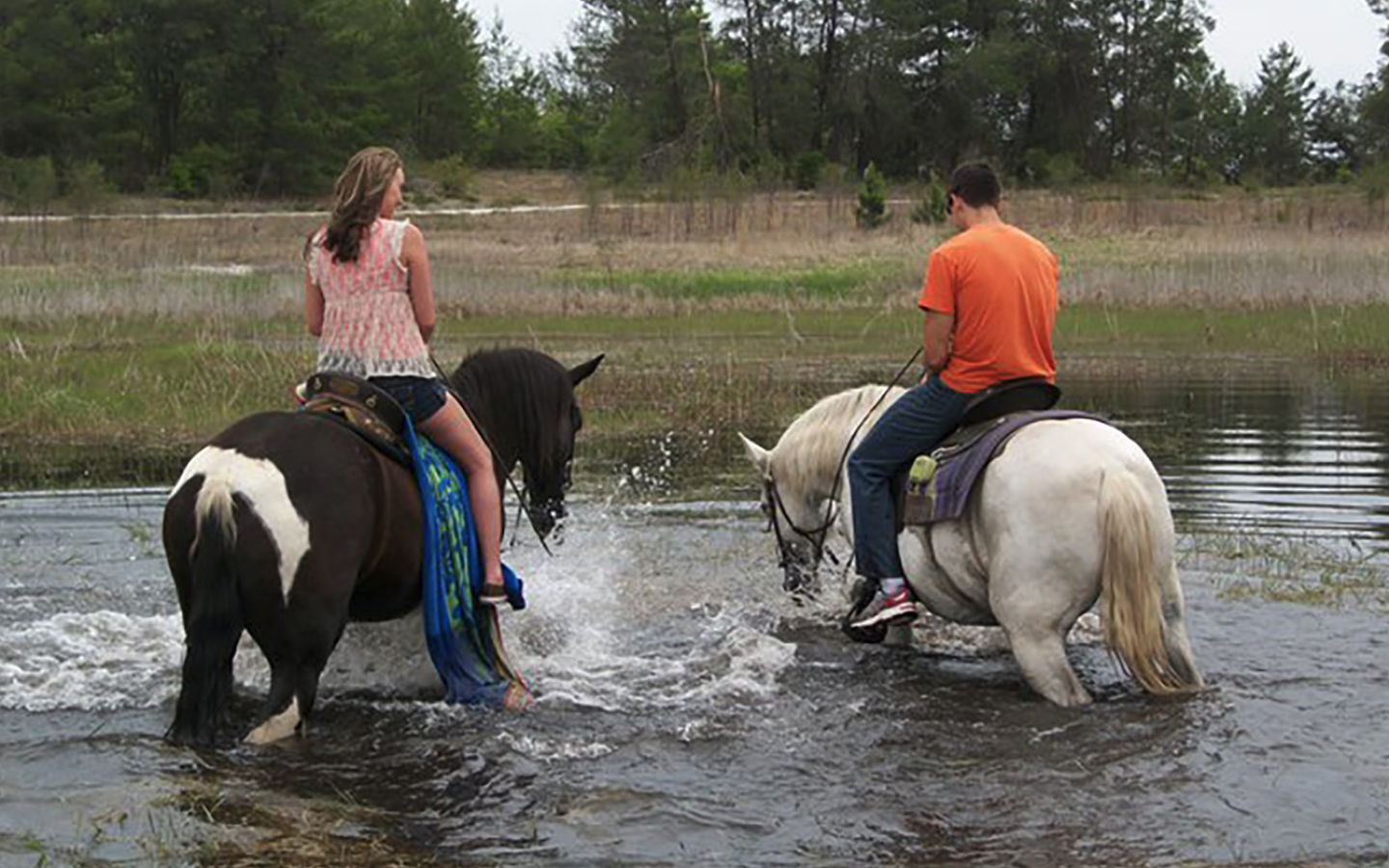 a man and woman, on individual horses, wading throw shallow water