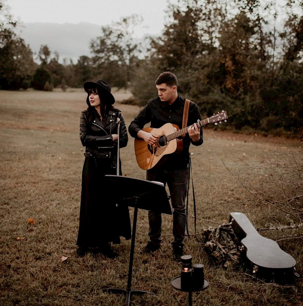 pat and jess standing in a field, with pat holding an acoustic guitar